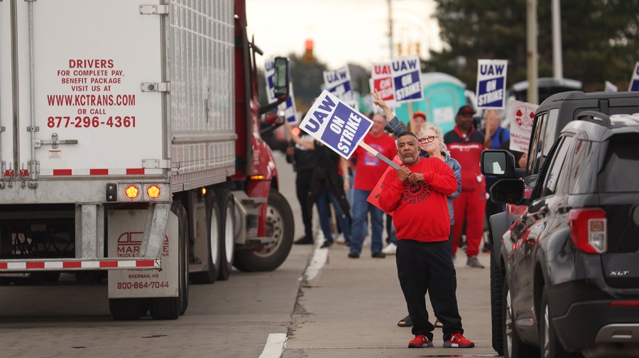 Workers hold blue and white "UAW on Strike!" signs next to a white cargo truck on a road