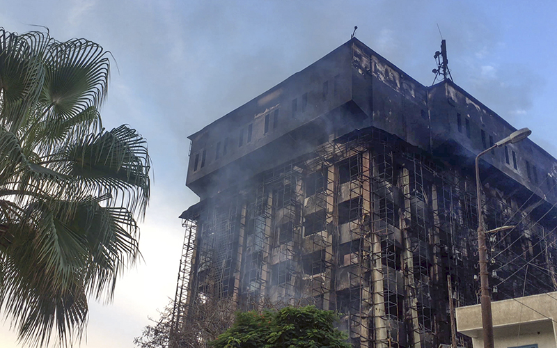 A burned building is seen covered with scorch marks. There is smoke rising from the building, and the windows are all broken.