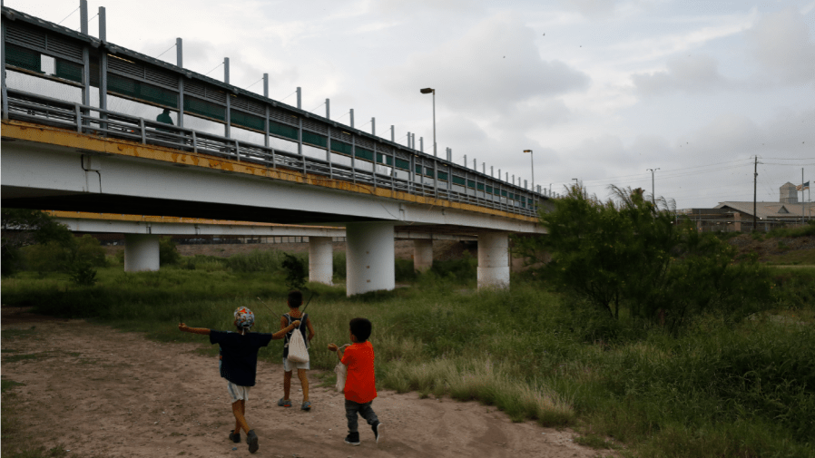 In this June 26, 2019 file photo, migrant children walk with their families along the Rio Grande, as pedestrian commuters use the Puerta Mexico bridge to enter Brownsville, Texas, seen from Matamoros, Tamaulipas state, Mexico.