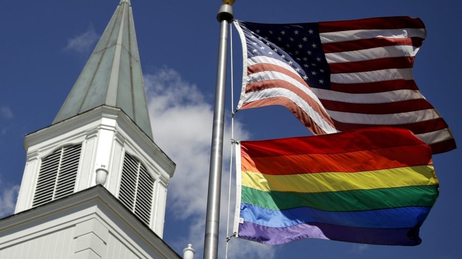 A gay pride flag flies along with the U.S. flag in front a church spire.