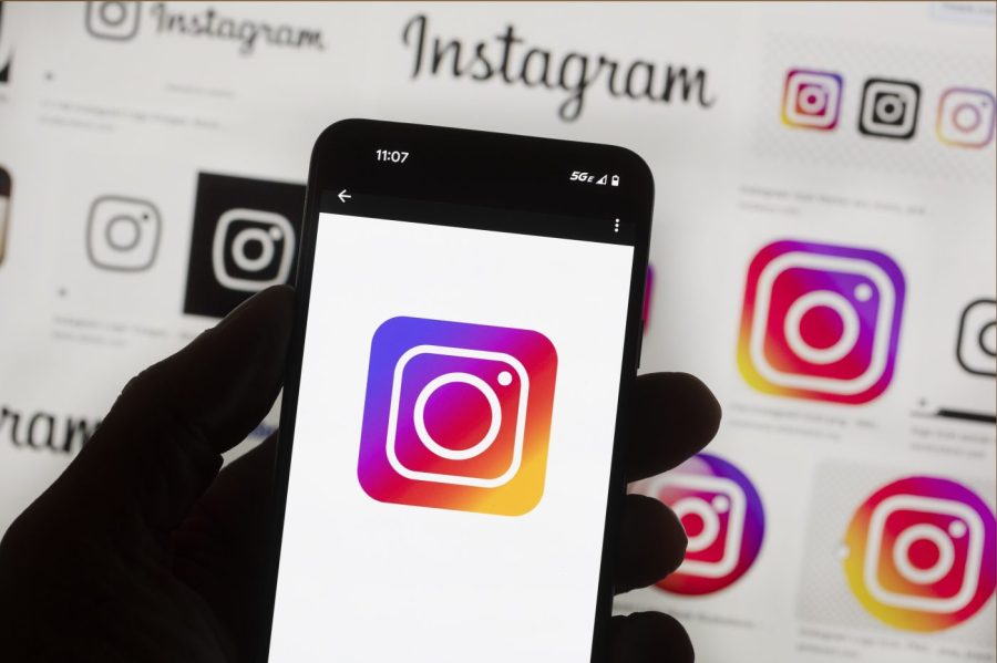 The Instagram logo is seen on a cell phone being held in front of a computer screen displaying more logos of the app.