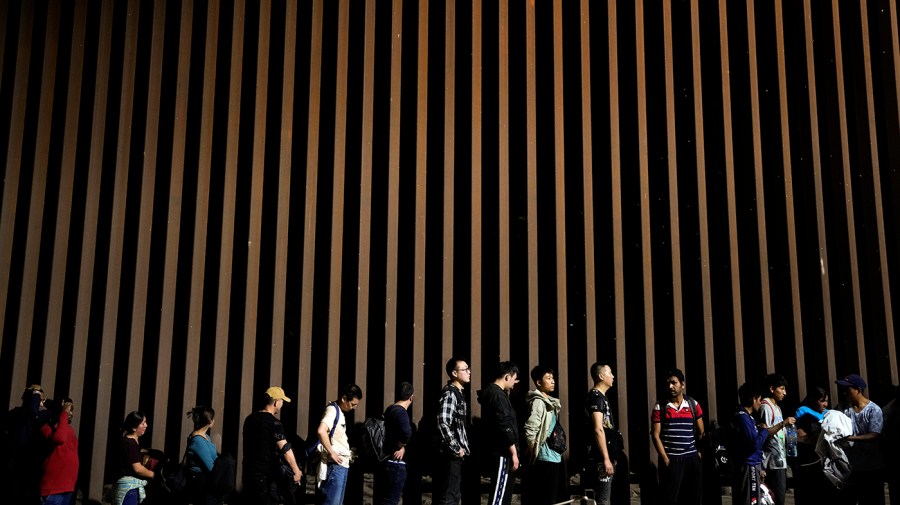 People line up against a border wall.