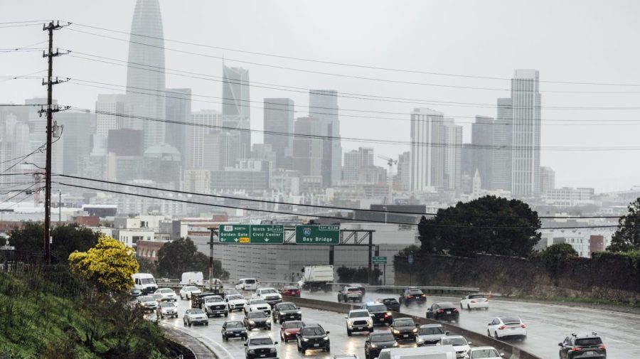 Motorists travel along a rain-soaked highway during a storm with the San Francisco skyline in the background.