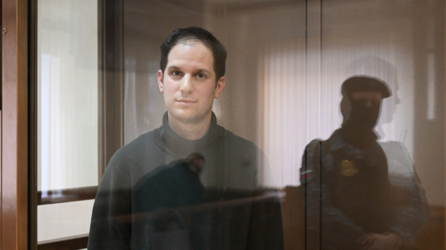 U.S. journalist Evan Gershkovich stands behind glass inside a defendants' room in a courthouse.