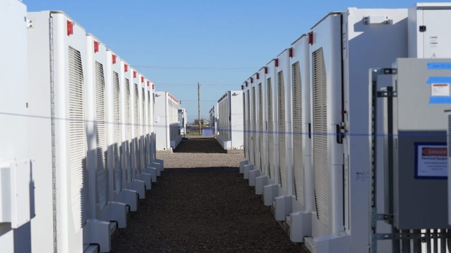 Rows of battery storage pods are seen outside against a blue sky.