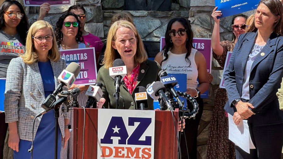 Arizona Attorney General Kris Mayes speaks to reporters as abortion-rights supporters hold signs in the background.