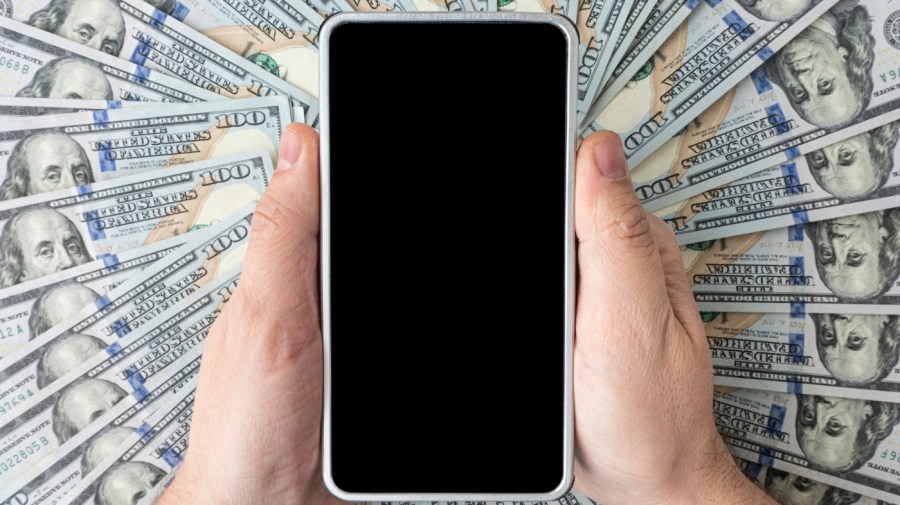 A person holds a blank phone over a backdrop of American bills.