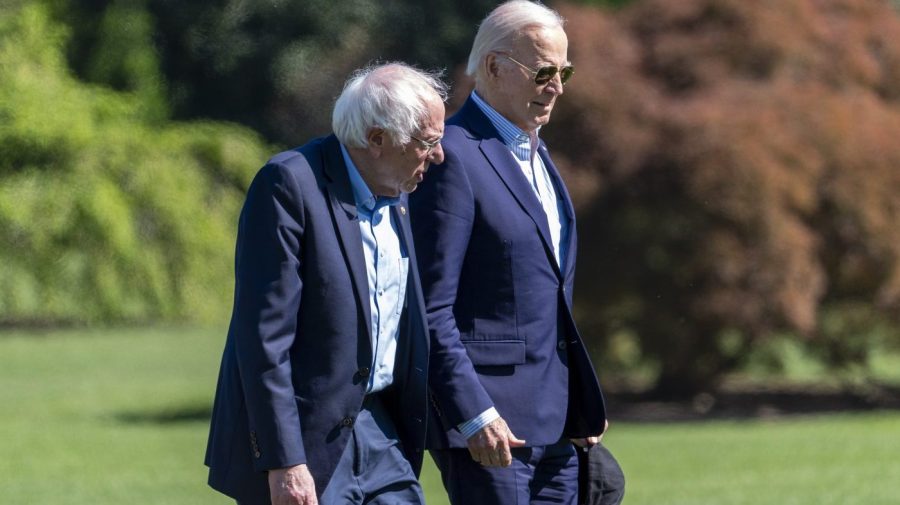 Senator Bernie Sanders and President Biden walk along a White House lawn.