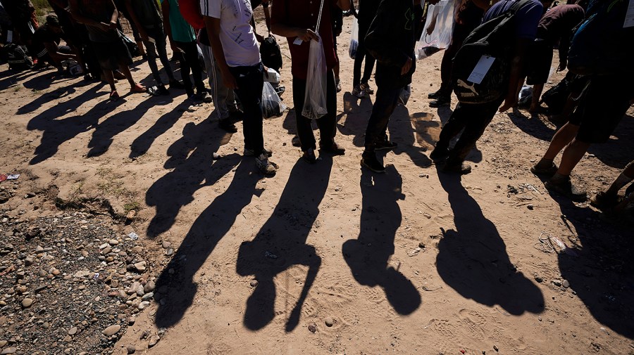 The shadows of migrants standing in line are visible on the ground.