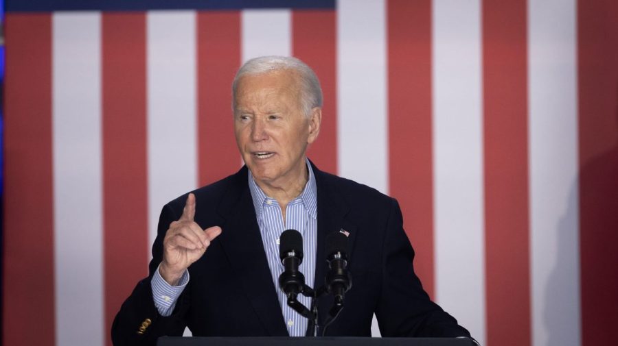 President Biden holds up a finger while speaking in front of large red-and-white stripes of an American flag.