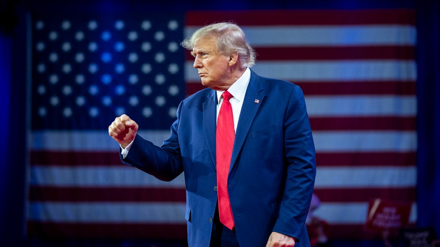 Former President Trump pumps his fist while on stage in front of a large American flag.