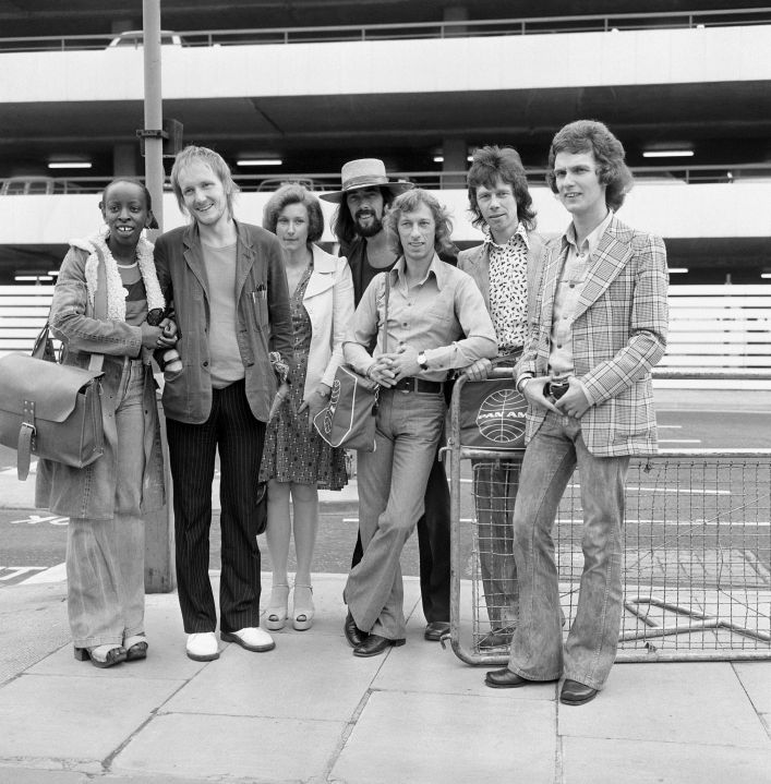 FILE - The Blue Mink pop group including Roger Cook, wearing hat, Madeline Bell, left, and bass player Herbie Flowers, second right, leaving London for Los Angeles, and the start of a two week tour on July 1973. Flowers, who played with David Bowie, Elton John, Lou Reed and other music legends in a decades-long career, has died at 86. (PA via AP, File)